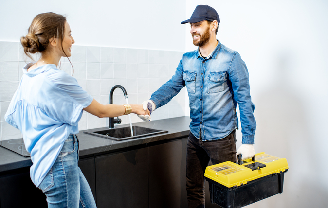 A professional handyman and a smiling homeowner shaking hands in a modern kitchen. The handyman holds a yellow toolbox, representing trust and satisfaction in home service interactions.