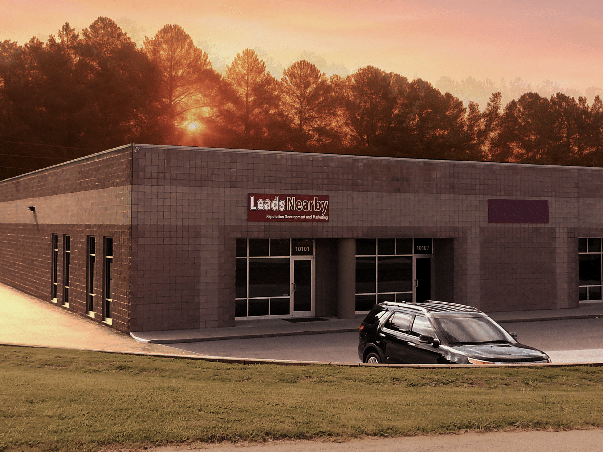 The sun rises over a one-story brick building with red LeadsNearby sign. Pine trees line the back of the building. Out front, a black SUV parked.
