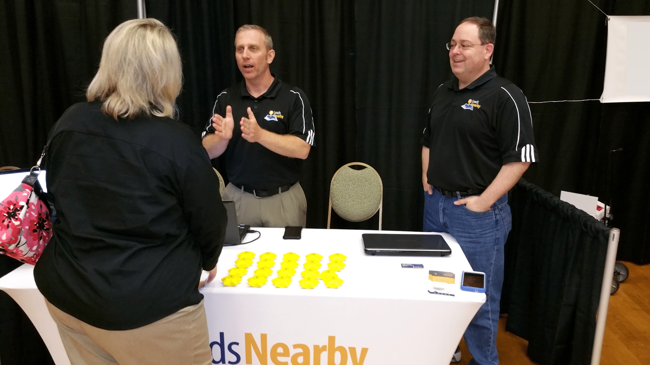 LeadsNearby booth at a tradeshow: Mark and Bob stand in black logo polos sharing home service contractor marketing lessons with woman; white table holds rows of yellow promotional stars, laptop, phones, black curtain backdrop.